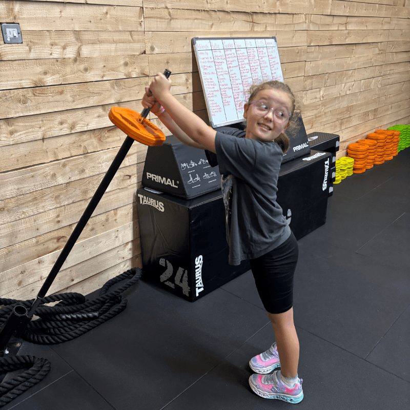 Young girl in a gym lifting a barbell with an orange weight plate, smiling near a wooden wall and whiteboard behind her.