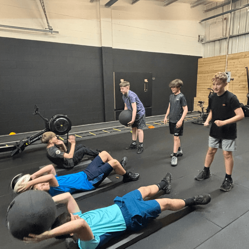 Group of boys in a gym doing core exercise on mats, holding medicine balls while others stand nearby and watch.