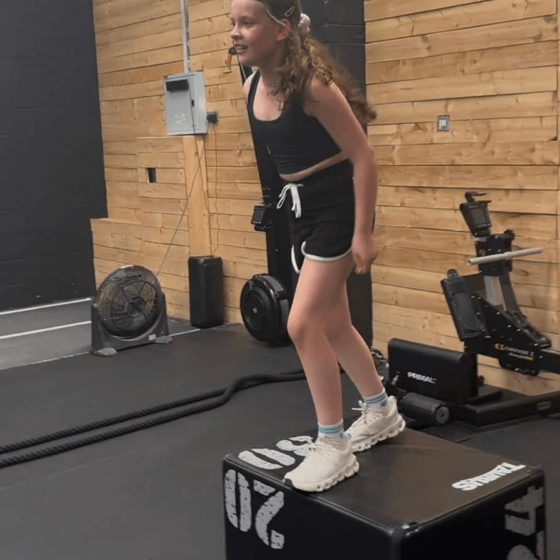 Girl in a black athletic outfit and headband stands on a plyo box, mid-exercise in a gym with wooden walls and equipment nearby.