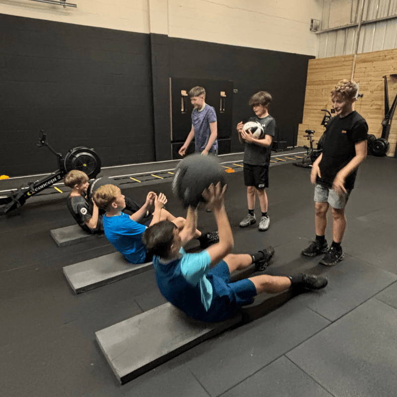 Group of boys doing sit-ups on inclined benches in a gym as a trainer and two peers watch nearby, one holding a medicine ball.