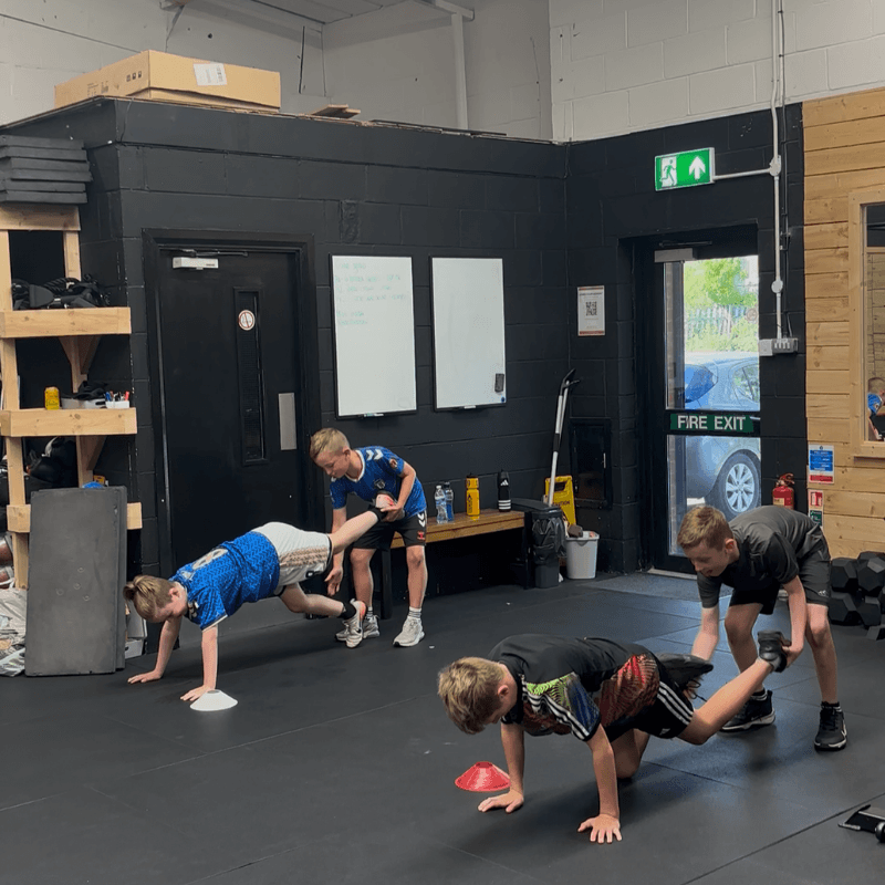 Group of four young men in a gym performing bodyweight exercises on a padded floor; one participant is being assisted by a trainer while others do push-ups near a red cone, with a black wall, whiteboards, and gym equipment in the background.