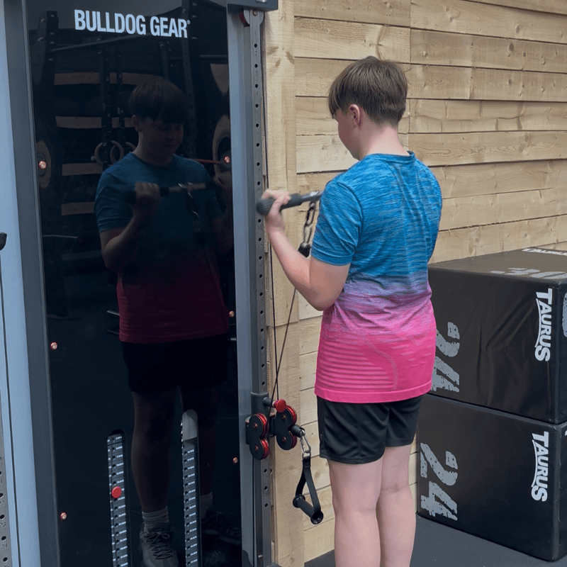 Boy in a blue-to-pink gradient shirt uses a cable machine, reflection visible in the glass door, gym setting with Bulldog Gear branding.