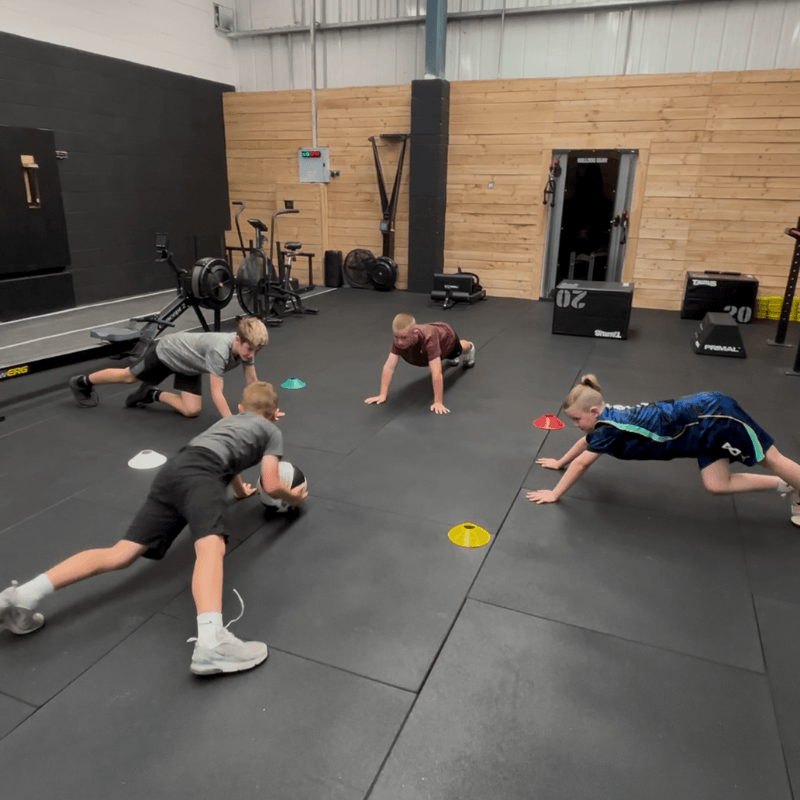 Four children in a gym doing bear crawls between colored cones on a padded floor.