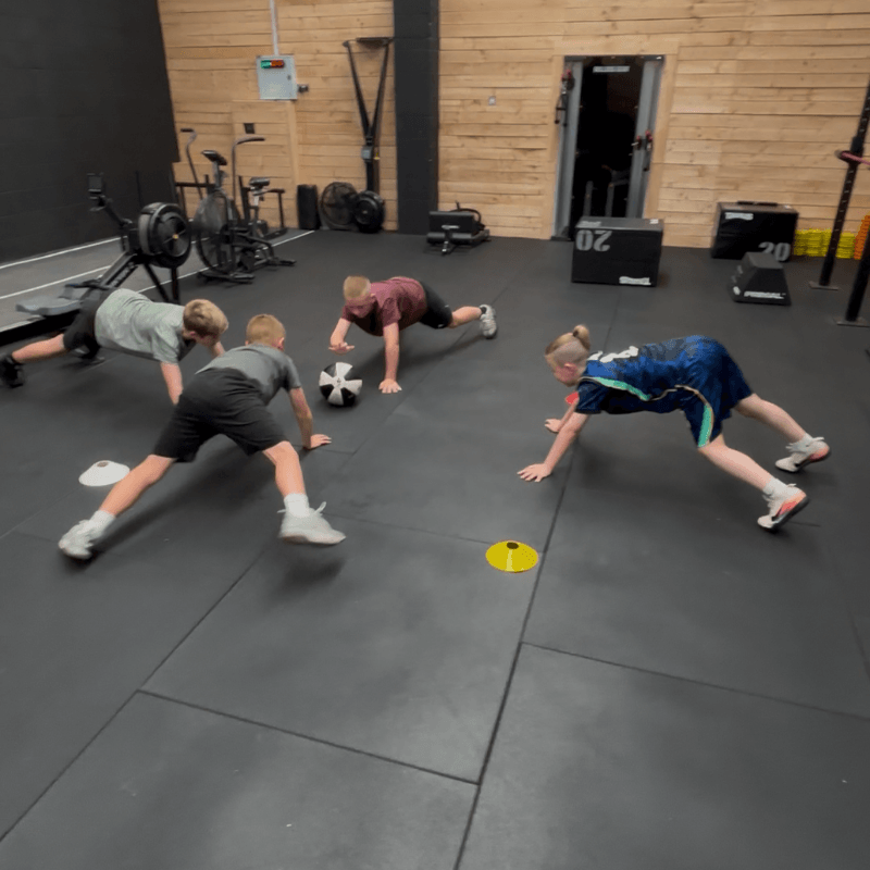 Four children in a gym doing bear crawls around a medicine ball on the floor near gym equipment.