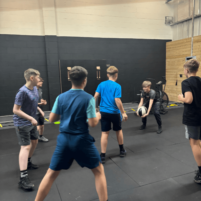 Group of boys playing a ball game in a gym, with black walls and padded mats nearby.