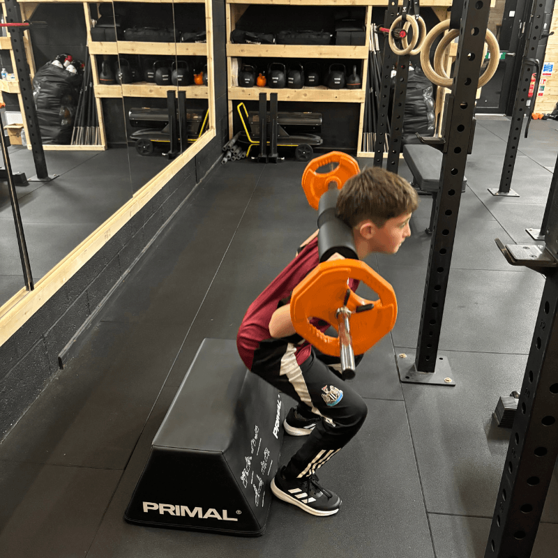 Young boy doing a squat on a padded block with an orange barbell and weight plates in a gym, wearing a maroon shirt and black pants.