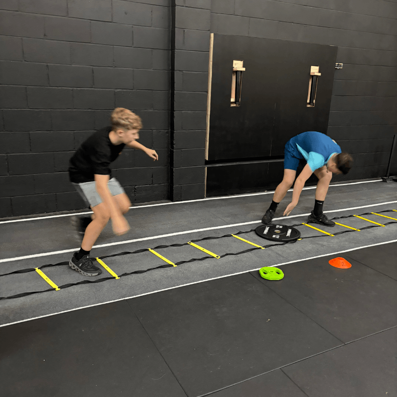 Two boys in a gym doing an agility ladder drill; one in a black shirt and gray shorts, the other in blue bending to grab a disk, with a yellow ladder on the floor.