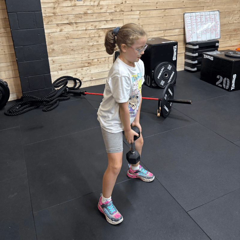 Young girl in a gym performing a dumbbell/kettlebell deadlift, knees slightly bent, gripping a weight between her legs.
