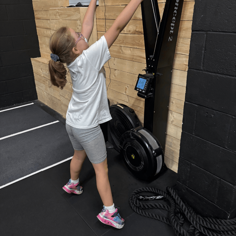 Girl in a white shirt and gray shorts reaches up to use a Concept2 ski erg in a gym, with a rope on the floor and a wooden wall behind.