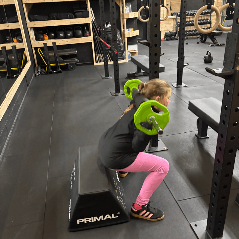 Girl squatting in a gym, holding bright green weight plates with a black plyo box in front and pink leggings/sneakers nearby