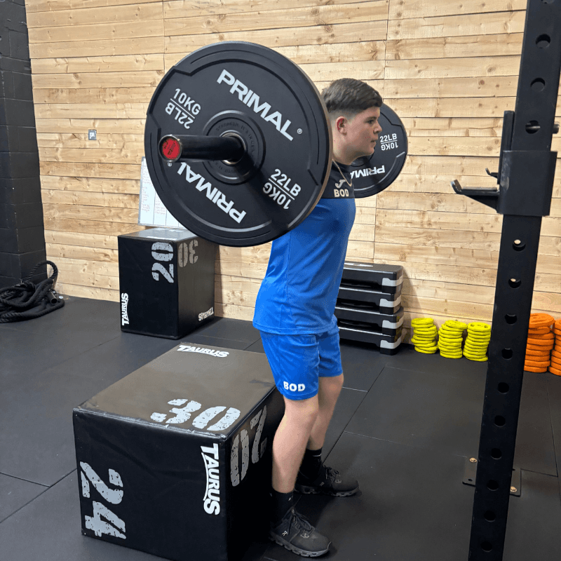 Man in blue shorts lifts a heavy barbell across his shoulders in a gym, preparing to squat or press.