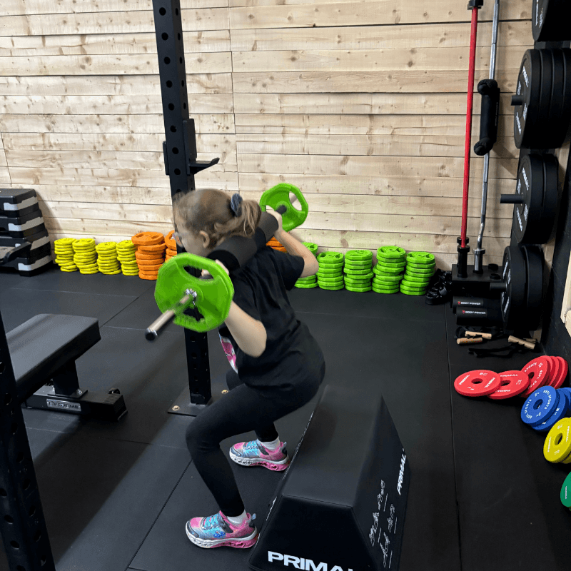 Person performing a squat with a green barbell on a black platform in a gym, with weight plates in the background