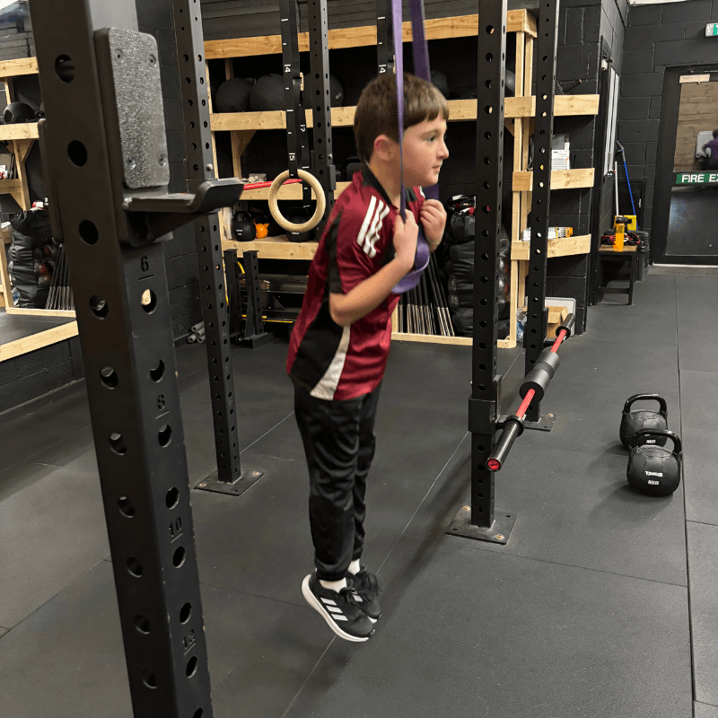 Young boy in a red shirt hanging from purple gym rings attached to a squat rack in a gym with kettlebells nearby.