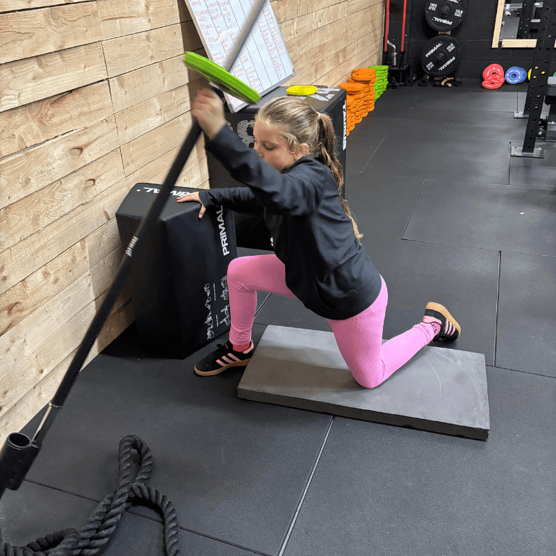 Young woman kneeling on a mat and pressing a landmine bar against a wooden wall in a gym.