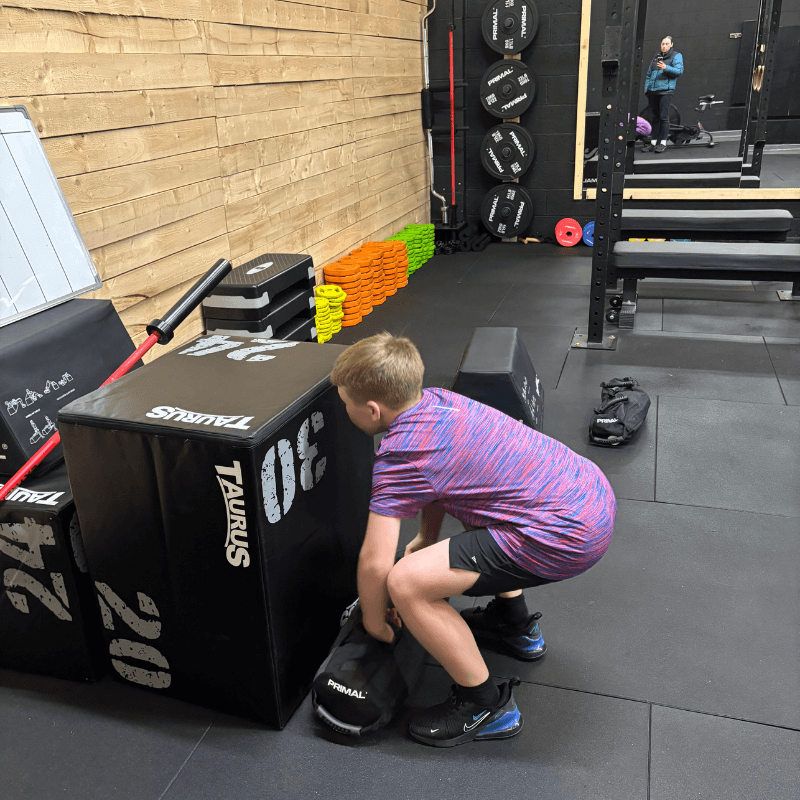 Young boy in a purple shirt squats to lift a large black Taurus box in a gym, with weights, colorful steps, and a person taking a photo in the mirror.