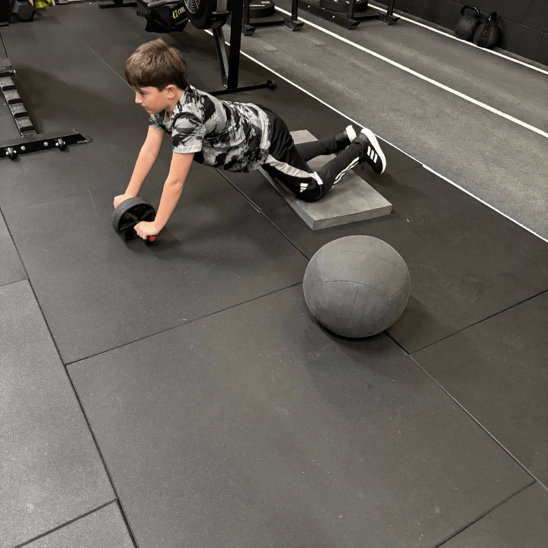 Boy in gym clothes performing an ab rollout on a padded mat, knees on a foam wedge with a gray medicine ball nearby in the foreground.