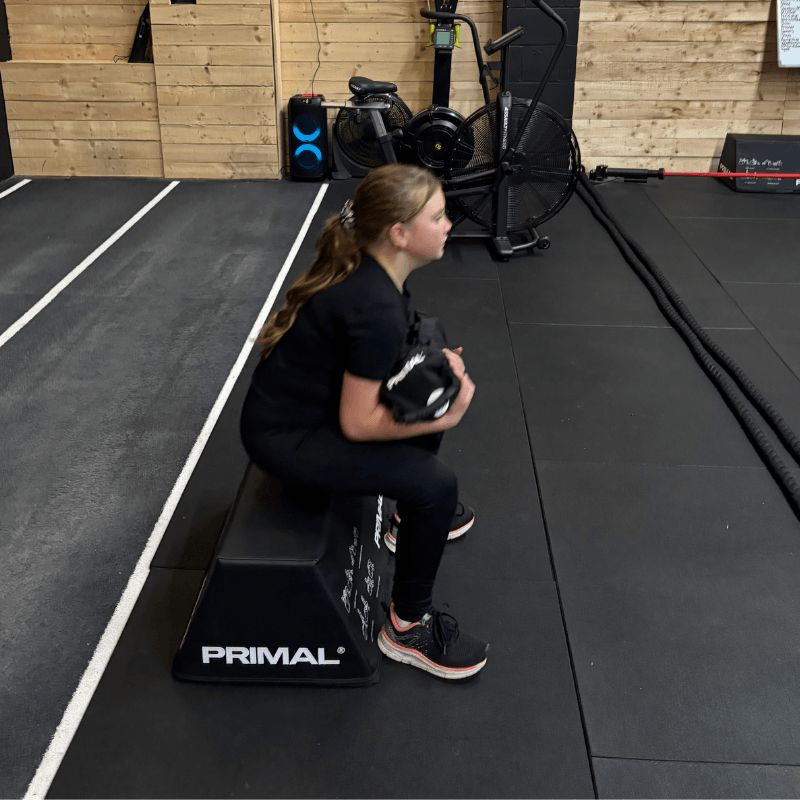 A young woman in a black outfit performs a squat holding a kettlebell at chest height on a black PRIMAL plyo box in a gym.