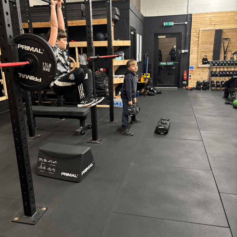 Young boy sits on a weight rack while another child stands nearby in a gym with lifting equipment and plates visible on the rack.