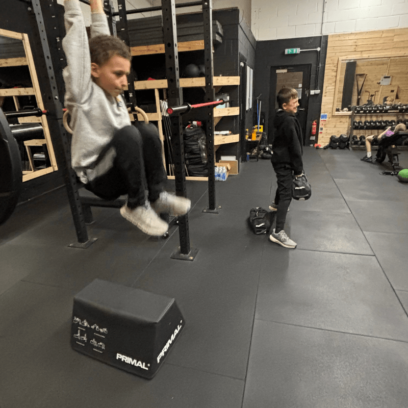 Two kids in a gym: one swinging on a low bar while another stands nearby with a bag and benches in the background. Note: This alt text should be concise and describe the action and setting rather than individual features.