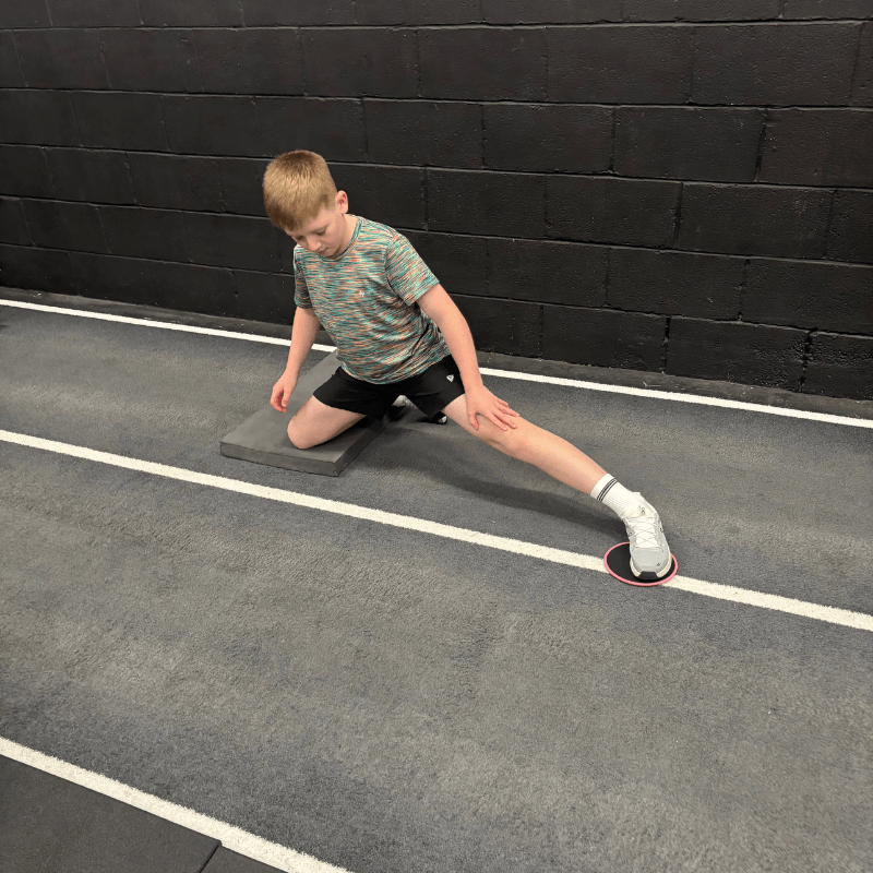 Boy in a striped shirt kneels on a mat in a gym, right leg extended to the side with a pink balance disc under the foot, performing a hip stretch.