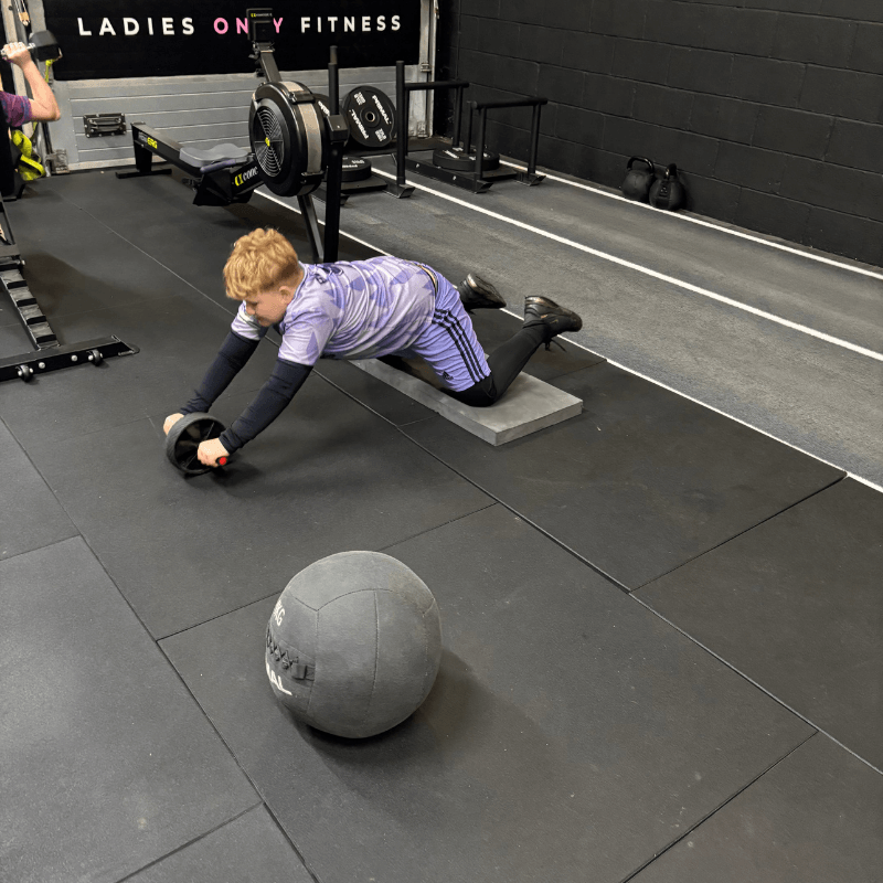 Person on hands and knees rolling a foam roller on a mat in a gym, with a large medicine ball nearby.