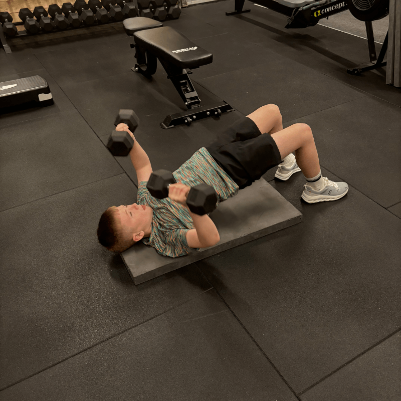 Person lying on a mat performing a dumbbell chest press in a gym, arms raised with dumbbells in each hand