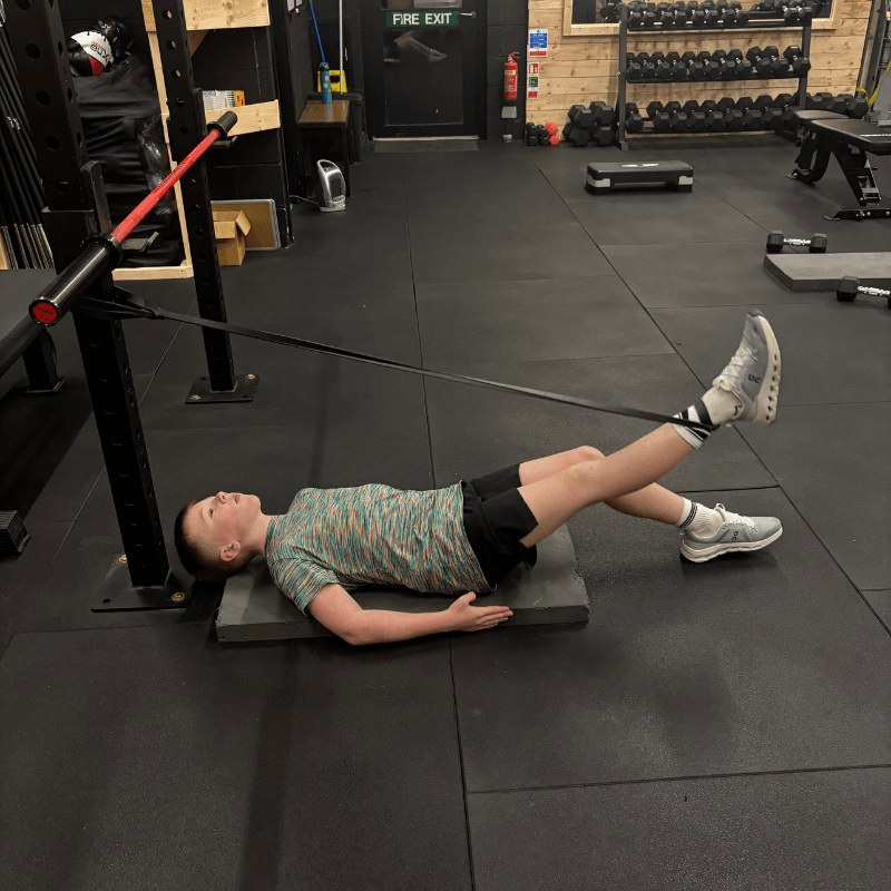 Person lying on a mat in a gym with legs raised and strapped to a resistance device attached to a squat rack, performing a leg drill.
