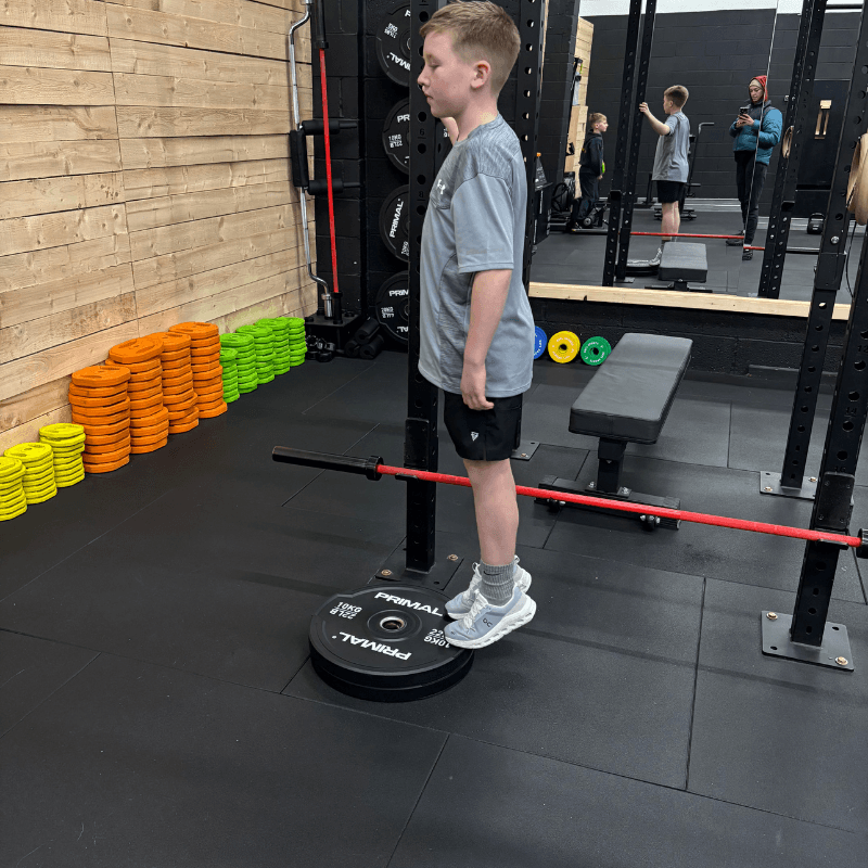 Boy in gray shirt and shorts stands on a weight plate next to a barbell on a squat rack in a gym, with colorfully stacked plates on the wall.