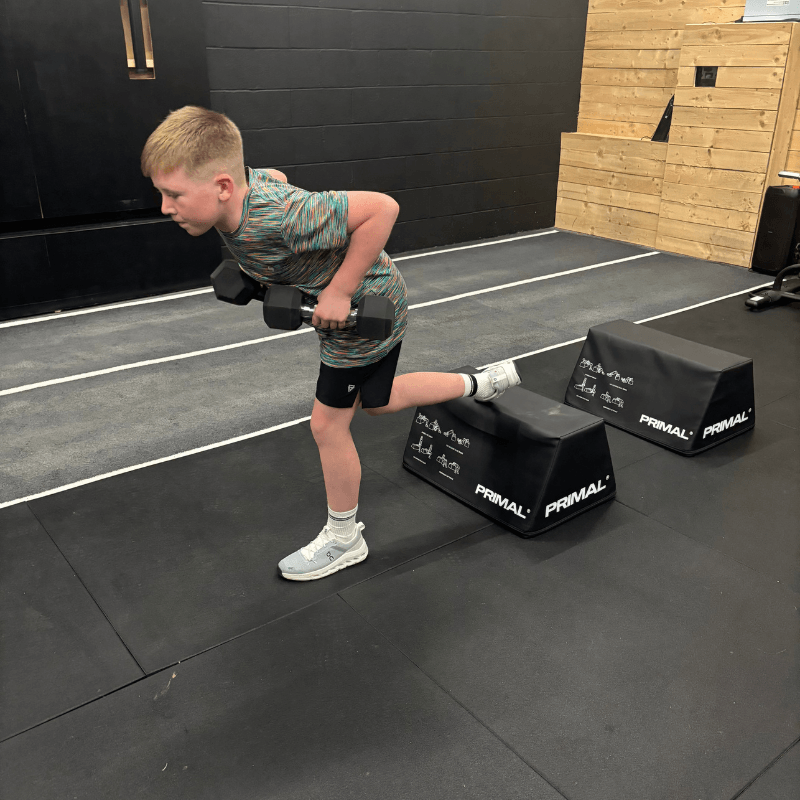 Young boy in a gym performing a bent-over dumbbell row with both dumbbells, one knee on a step platform for balance