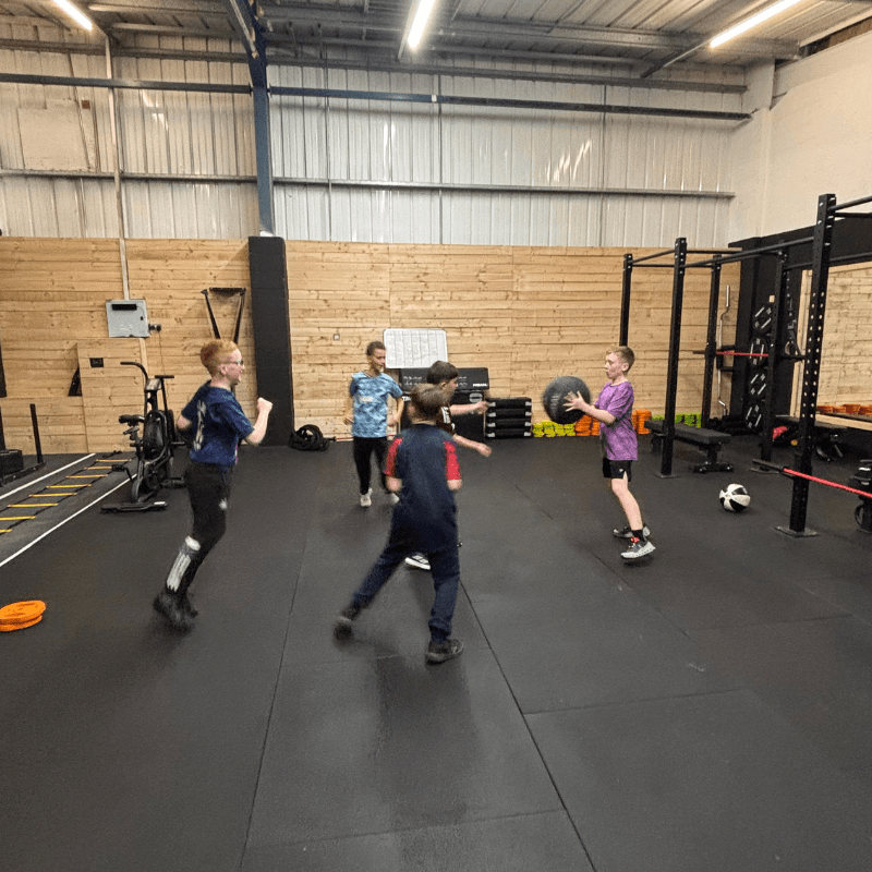 Five children in a gym playing with a medicine ball on a black padded floor