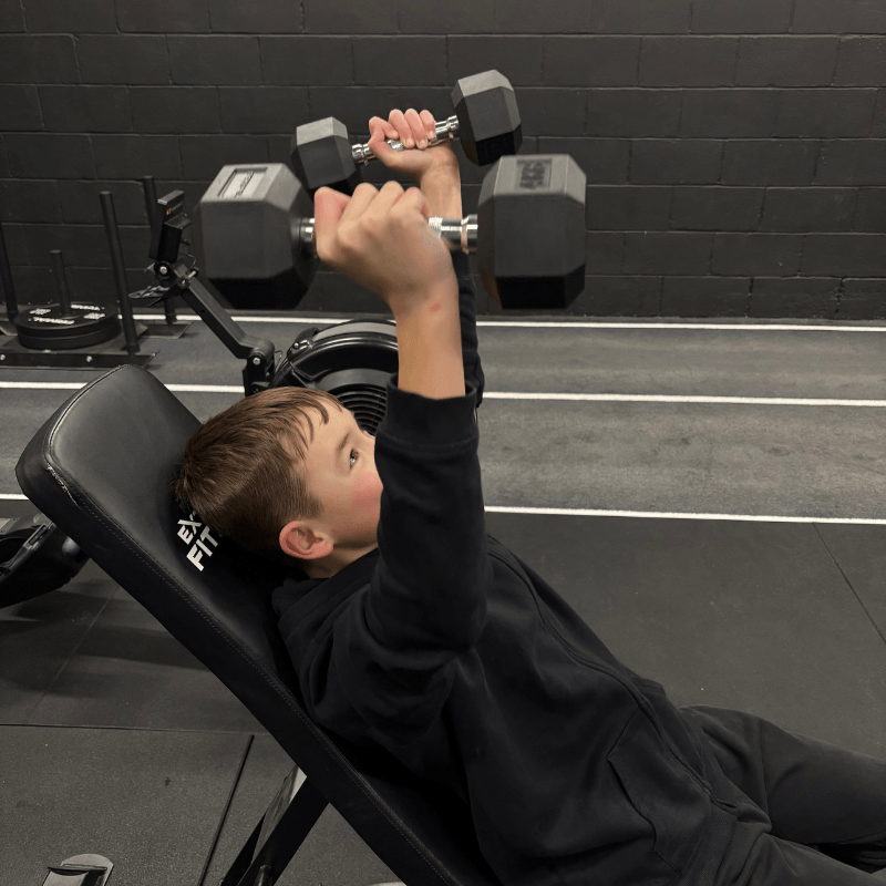 Boy wearing a black hoodie lies on an incline gym bench and lifts two dumbbells overhead in a weight room.