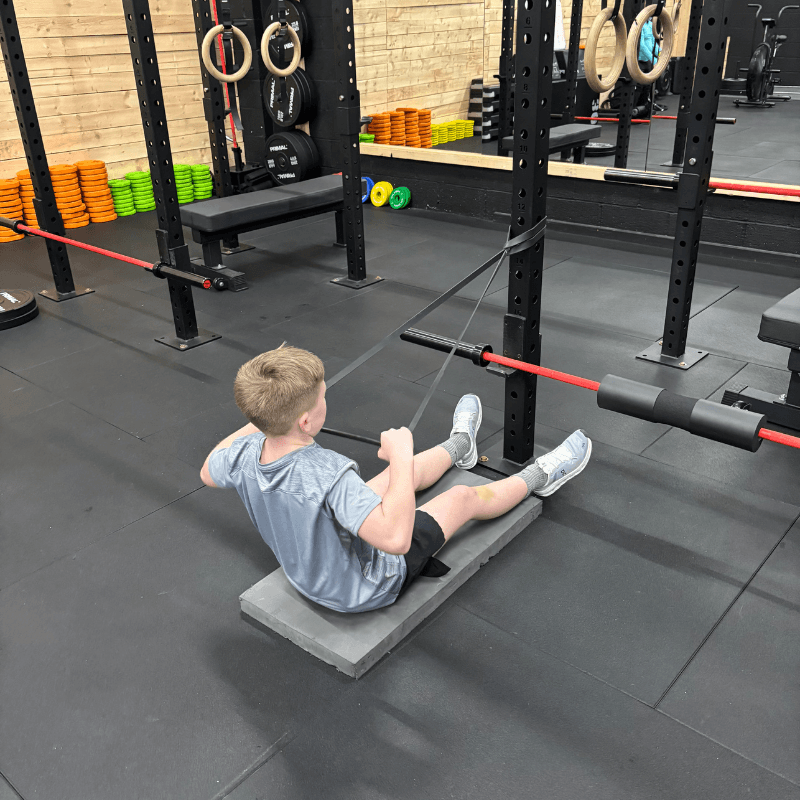 Young man in a gym doing a floor row with resistance bands attached to a squat rack.