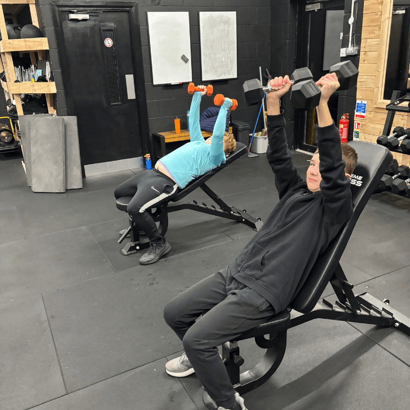 Two people in a gym performing dumbbell presses on incline benches, one in the foreground and one in the background.