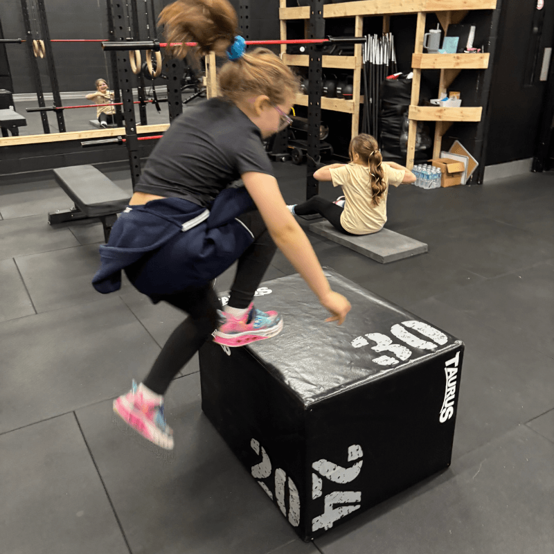 Young girl mid-jump over a black plyometric box labeled Taurus; another child sits on a mat in the background in a gym.