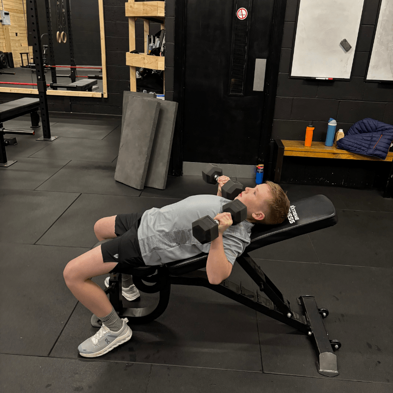 Man lying on an adjustable bench, performing dumbbell bench press with two 25–30 lb dumbbells in a gym.”,