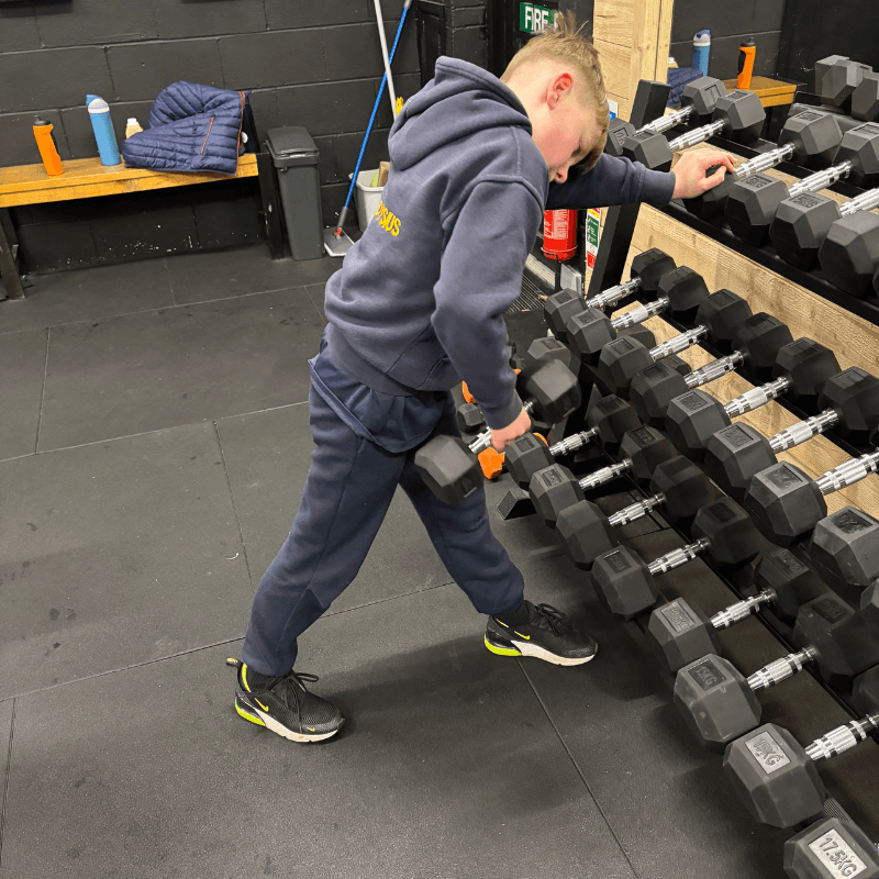 Young person in a navy tracksuit selects a dumbbell from a rack in a gym.
