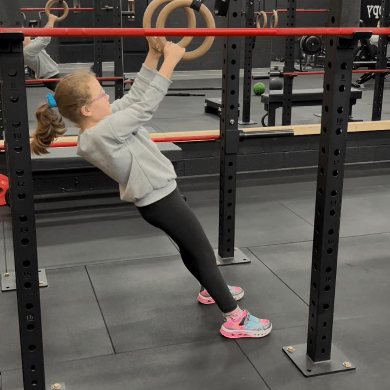 Young girl in a gray hoodie and black leggings performs ring rows, pulling up on gymnastic rings mounted to a power rack in a gym.