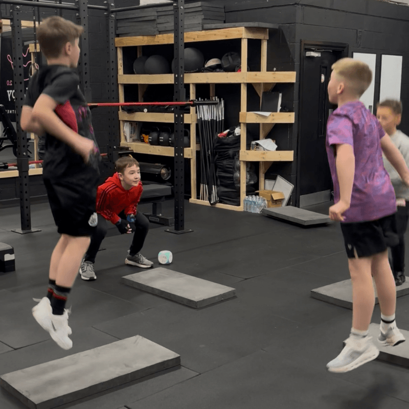 Group of children jumping over elevated platforms in a gym obstacle course, with weight racks and equipment in the background.