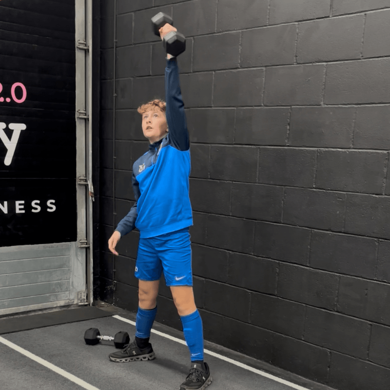 Person in blue athletic gear lifting a dumbbell overhead in a gym corner