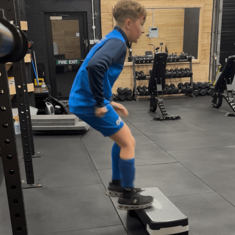 Young person in blue athletic gear performing a box jump on a small platform in a gym area with weights in the background.