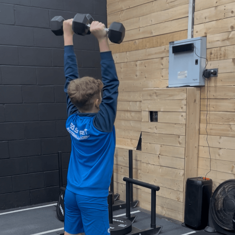 Person in blue athletic wear lifts two dumbbells overhead in a gym with black and wooden walls.