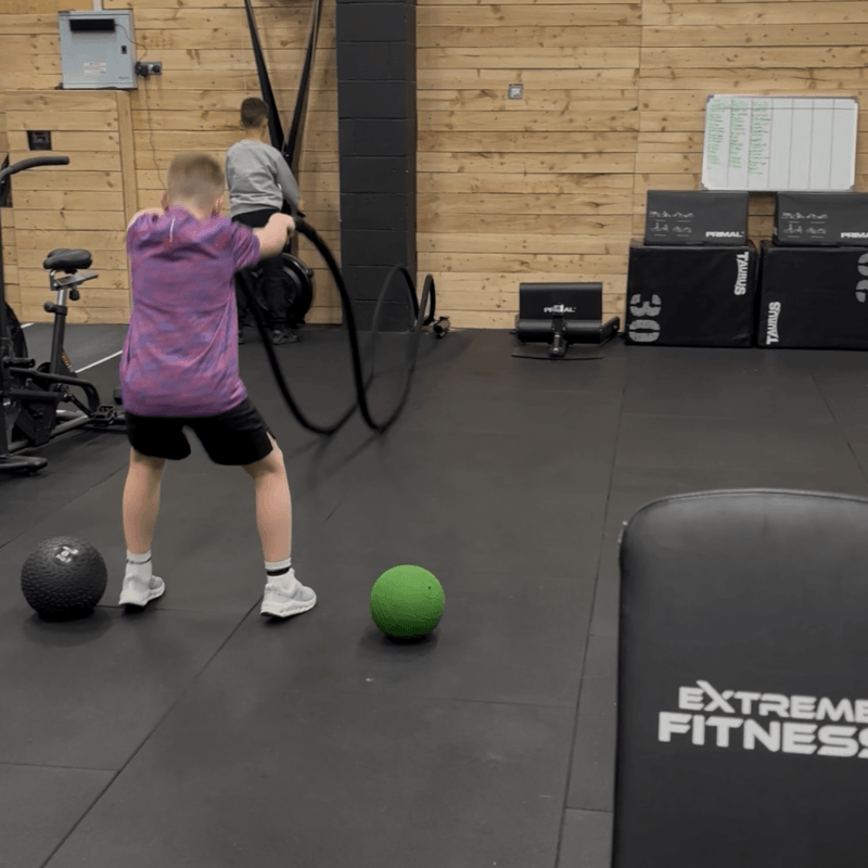 Two children exercising in a gym: one swinging a battle rope while the other faces a black wall; a green medicine ball and a textured black ball sit on the floor nearby.