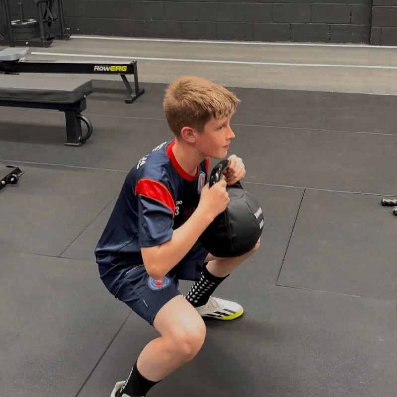 Young boy in a sports uniform crouches on gym mats while holding a black medicine ball near his chest for support during training cool-down or drill.