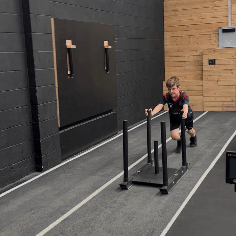 Athlete in a gym pushing a weighted sled on an indoor track, performing a push exercise toward the camera.