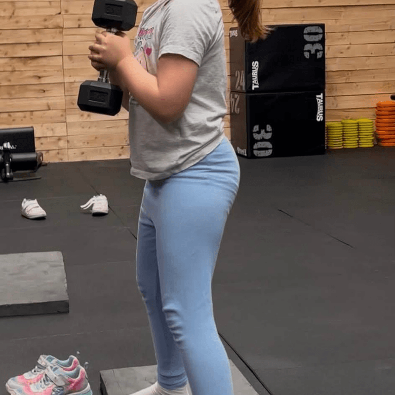 Person in a gym holding a dumbbell with both hands at chest height, wearing a gray shirt and light blue leggings.