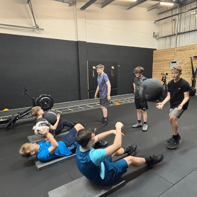 Group of boys in a gym doing sit-ups on mats while others hold a medicine ball nearby, with gym equipment and a black wall in the background.