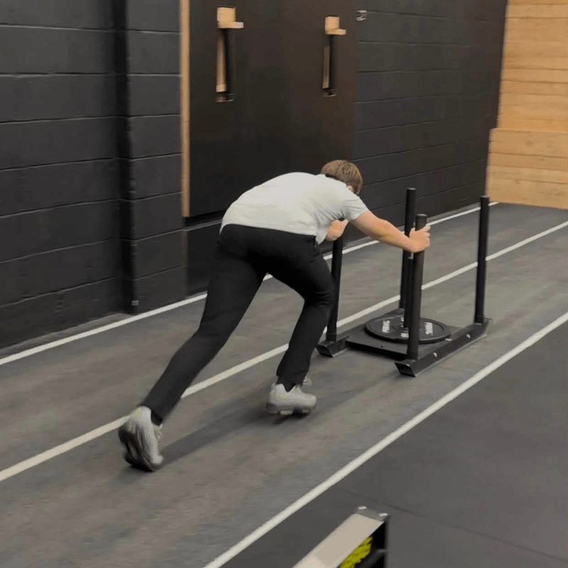 Man in a grey shirt and black pants pushing a weighted sled along a gym floor for sprint training.