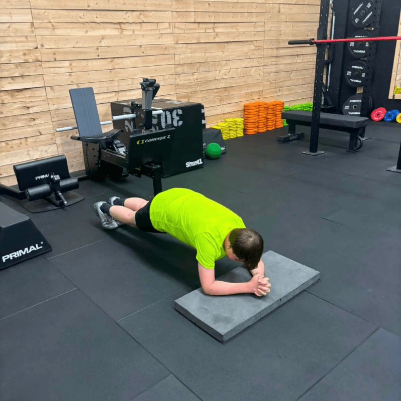Person in a neon shirt holds a forearm plank on a gray mat inside a gym, with weights and equipment in the background.