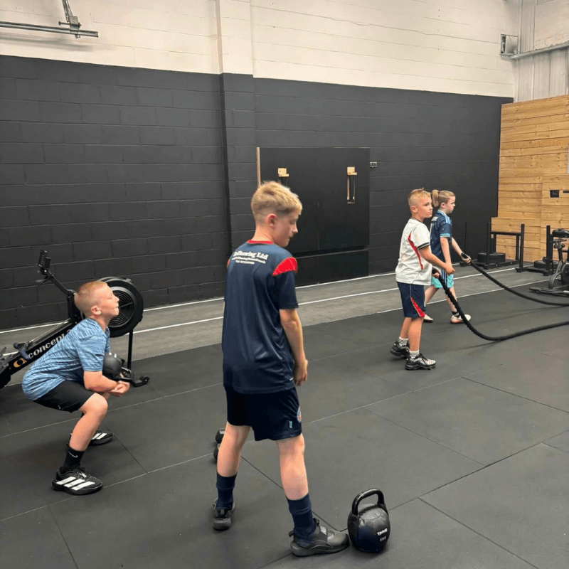 Group of young boys in a gym performing conditioning with battle ropes and kettlebell on a black rubber floor.