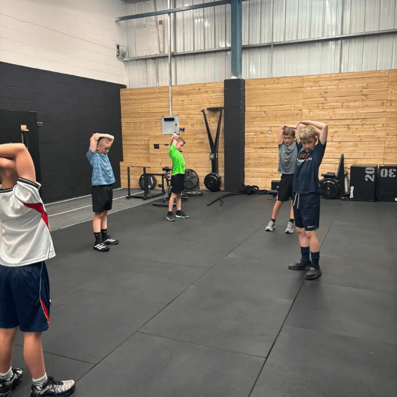 Group of young boys in a gym standing apart, hands behind their heads, doing a standing stretch on a padded floor with equipment in the background.